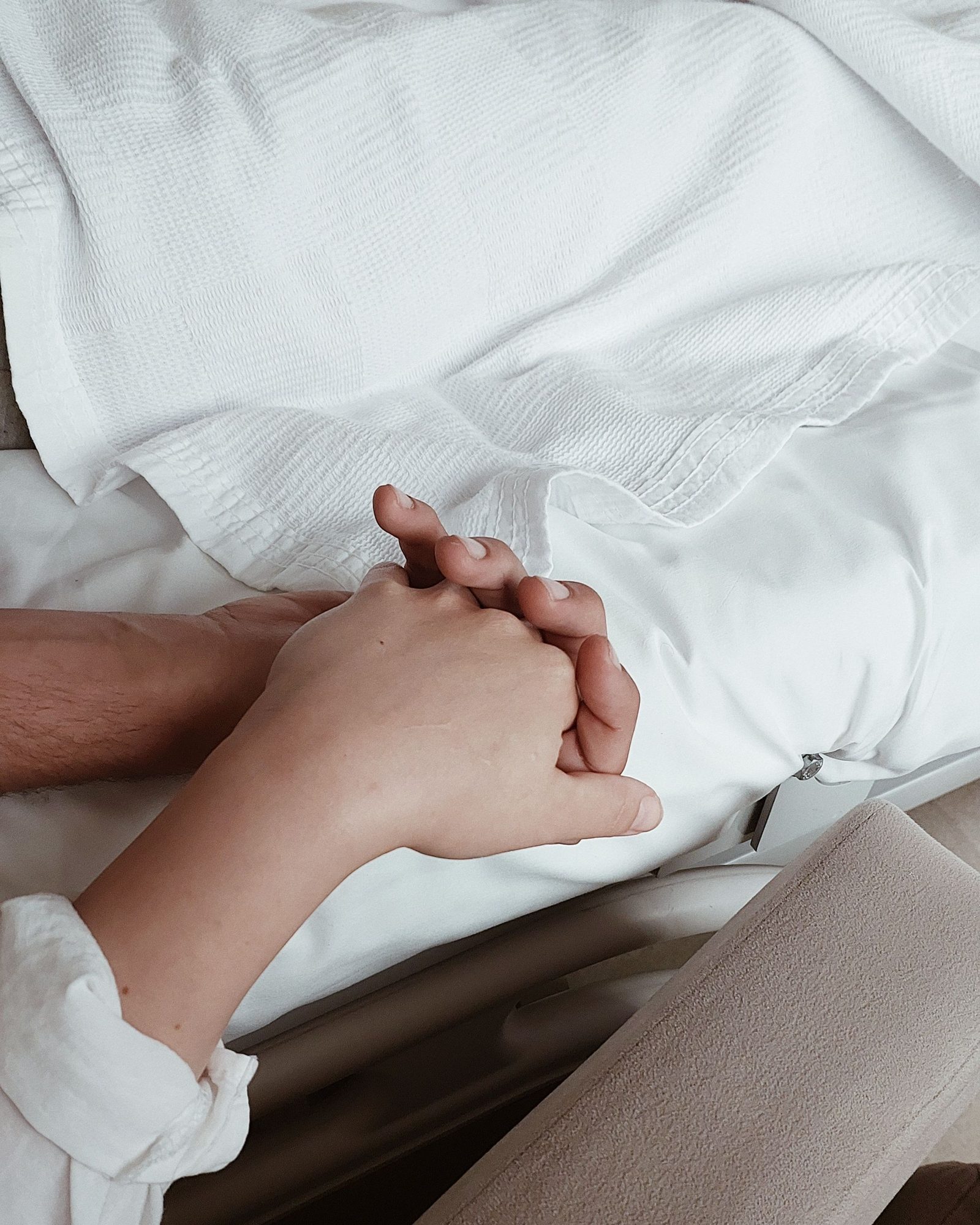 Two people's hands clasped on a bed
